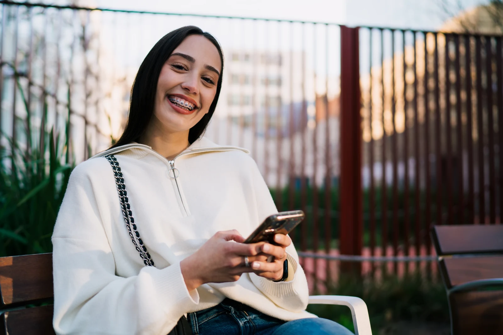A smiling patient with braces holding a phone outdoors